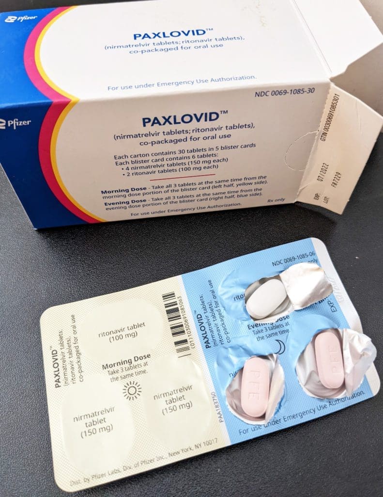 A box of Paxlovid and an opened blister pack showing pink oval tablets and white oval tablets, both labeled with dosages. The packaging includes usage instructions and emergency use authorization details.