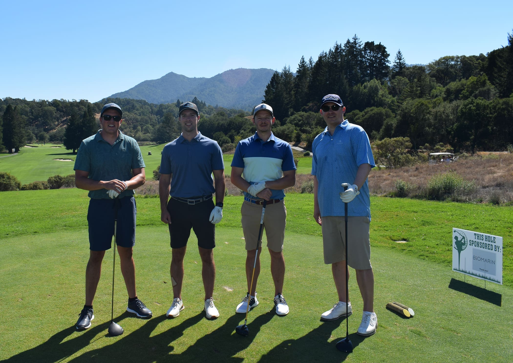 Four men in golf attire stand on a sunny golf course with clubs in hand. Trees, hills, and a clear blue sky are in the background. A sponsor sign is visible on the right side of the image.
