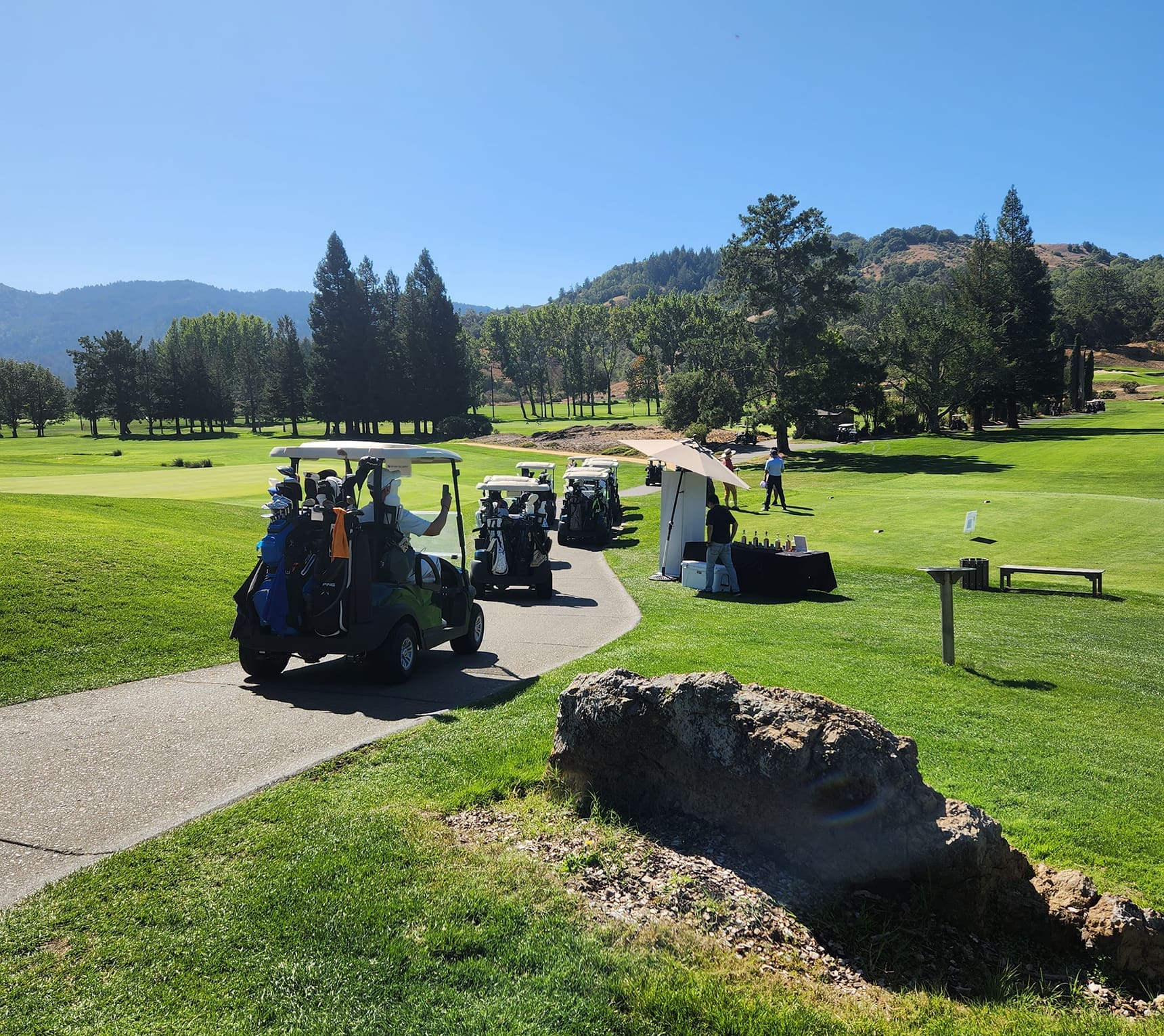 Golf carts are parked on a paved path next to a table with drinks on a sunny golf course. Trees, hills, and clear blue sky are in the background. A person is standing near the table.