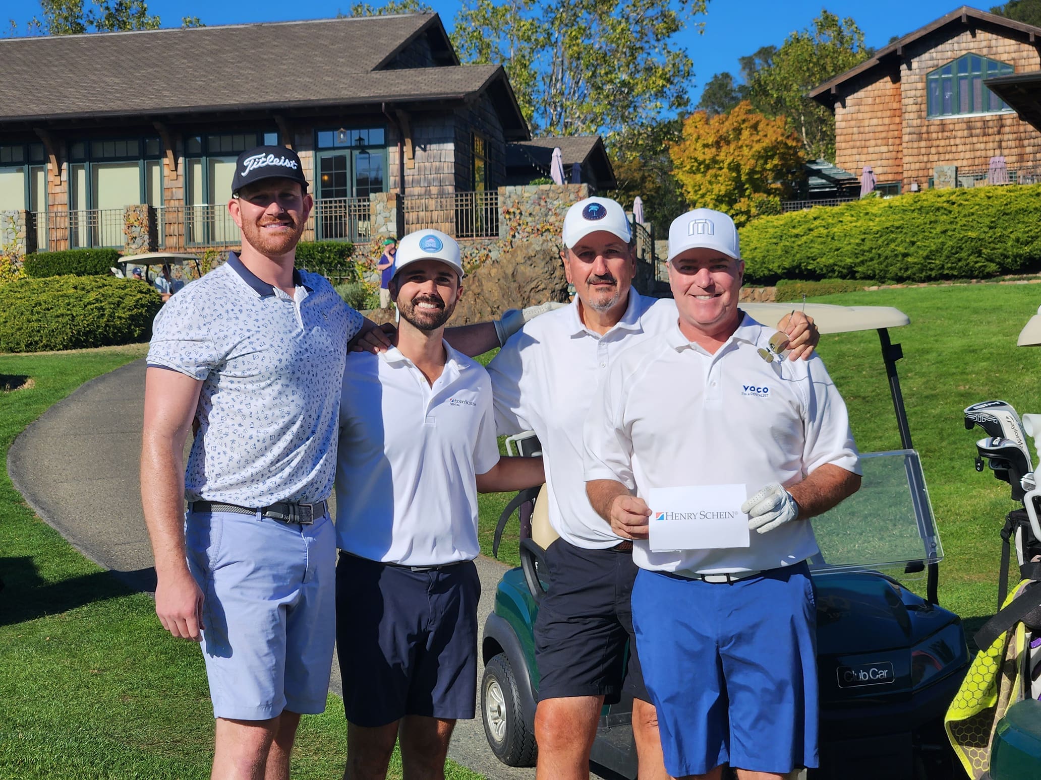 Four men pose together on a golf course next to a golf cart, all dressed in golf attire and hats, with lush green grass and a clubhouse in the background. One man holds a paper that says Stinson Scott.