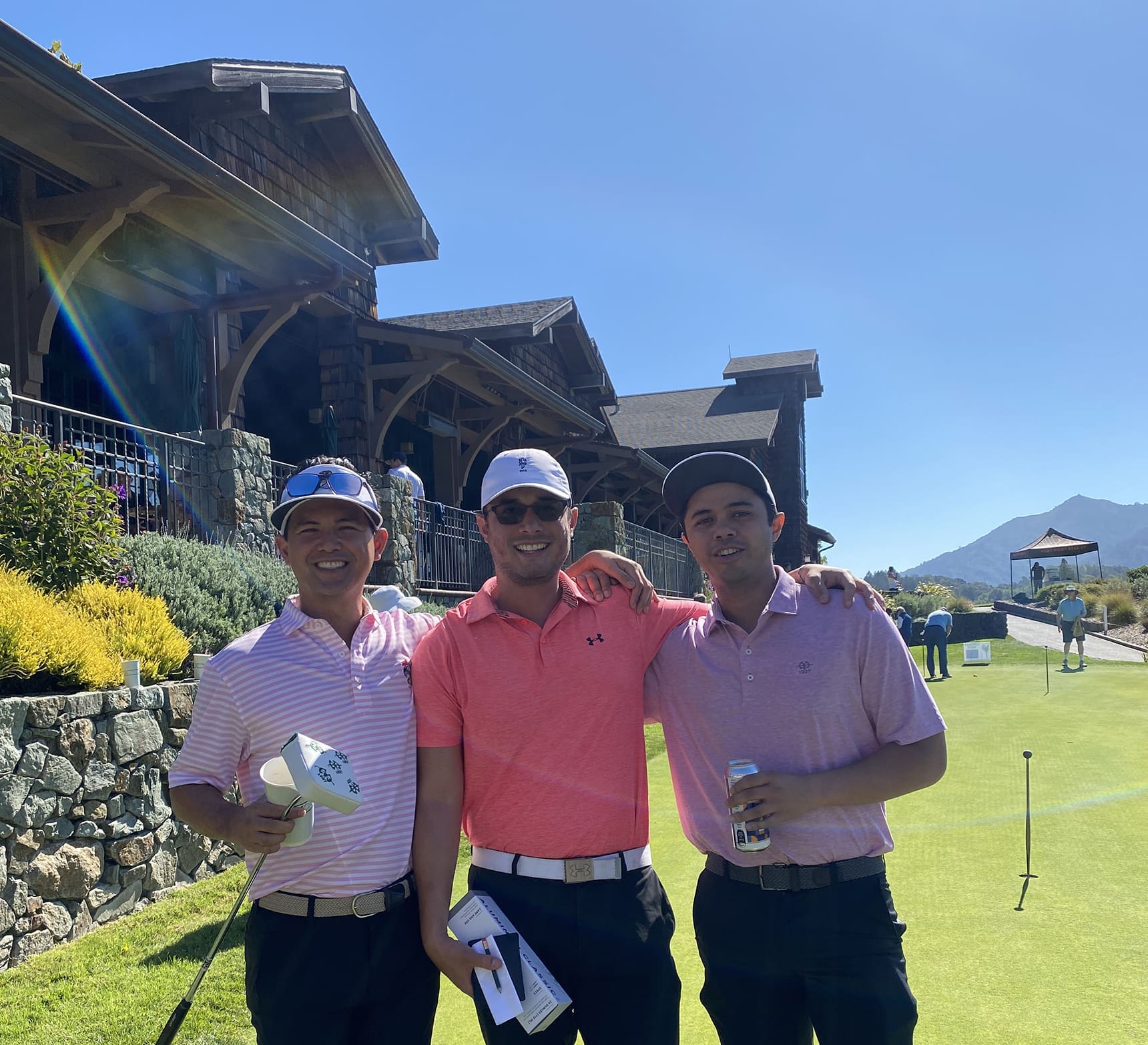 Three men in golf attire, smiling with their arms around each other, stand on a golf course near a clubhouse on a sunny day. They are wearing sunglasses and holding drinks and hats. The background shows hills and blue sky.