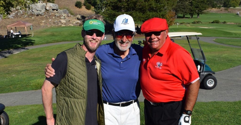 Three smiling men stand arm-in-arm on a sunny golf course, wearing golf attire and hats. A golf cart and green fairway are visible in the background.