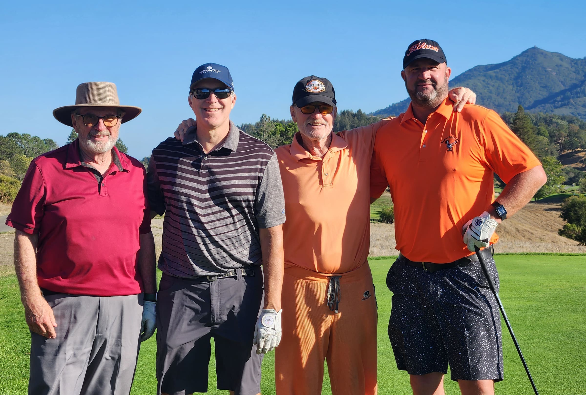 Four men stand together on a golf course, smiling at the camera. They wear golf attire, with mountains and trees visible in the background under a clear blue sky.