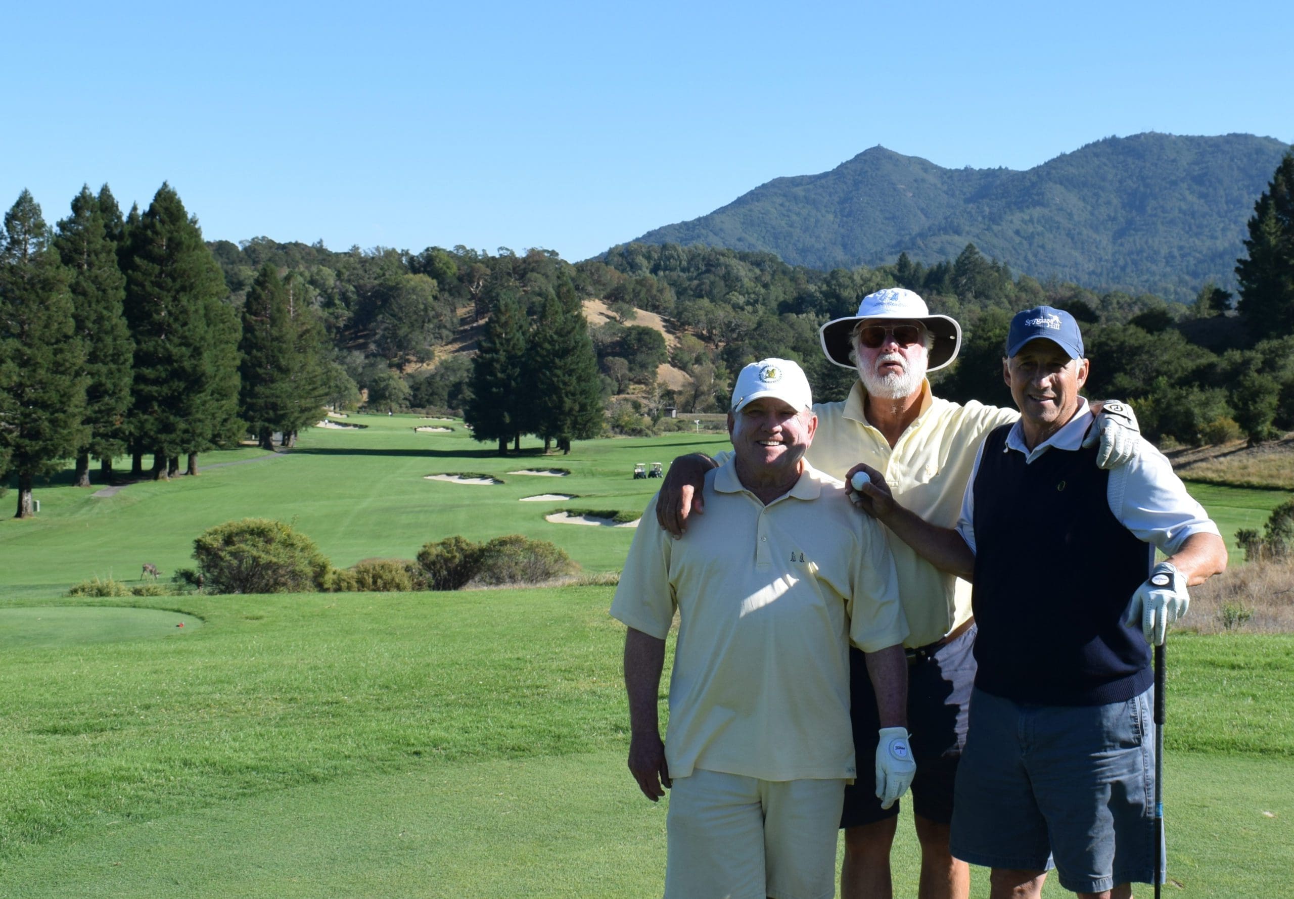 Three men in golf attire stand smiling on a sunny golf course with green grass, sand bunkers, tall trees, and mountains in the background.