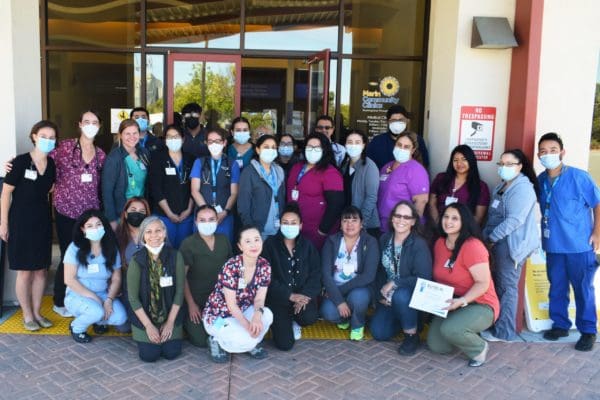 A large group of healthcare workers, many wearing masks and scrubs, pose and smile outside a medical facility entrance. Some individuals kneel in front while others stand behind them. One person holds a certificate.
