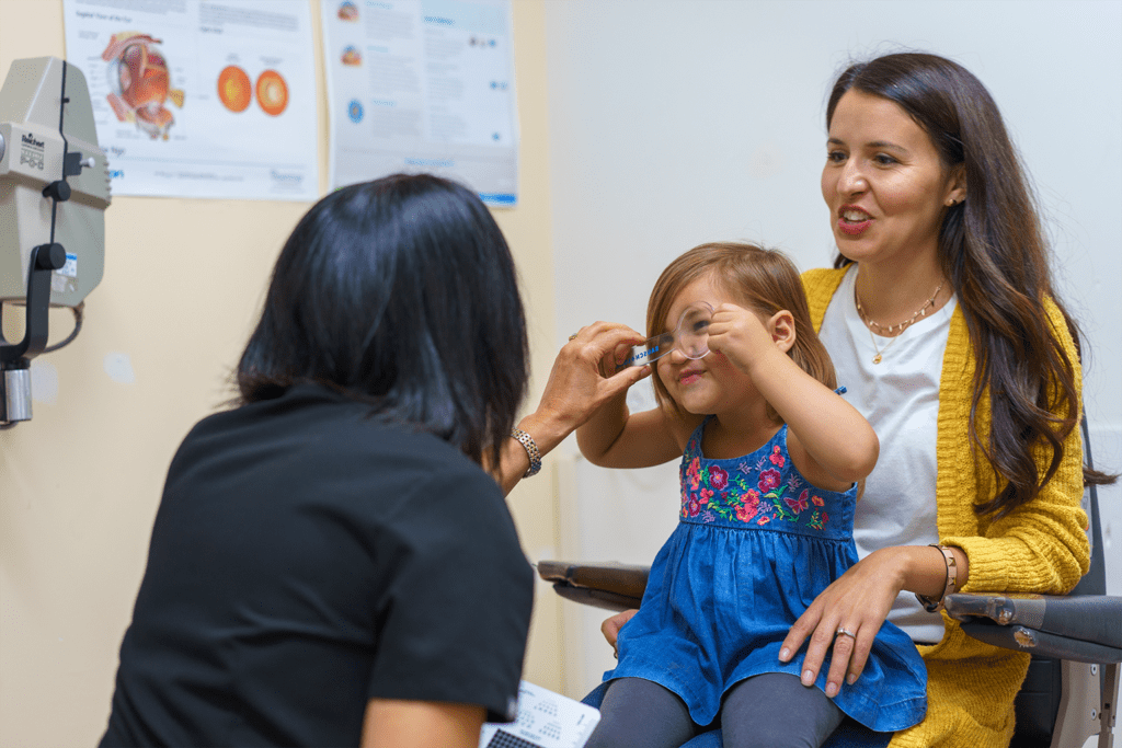 A young girl sits on a woman’s lap during an eye exam while a healthcare professional holds up a lens for her to look through. Eye health posters are visible on the wall in the background.