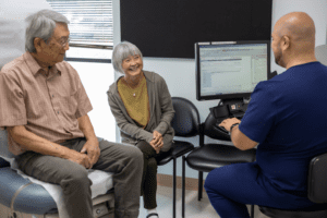 An older man sits on an exam table, with an older woman smiling beside him, while a healthcare professional in blue scrubs types on a computer during a medical consultation in a clinic room.
