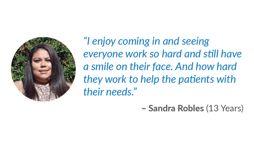 A woman with long dark hair smiles outdoors. Next to her is a quote about enjoying seeing hardworking, smiling colleagues who help patients, attributed to Sandra Robles, with 13 Years noted in parentheses.