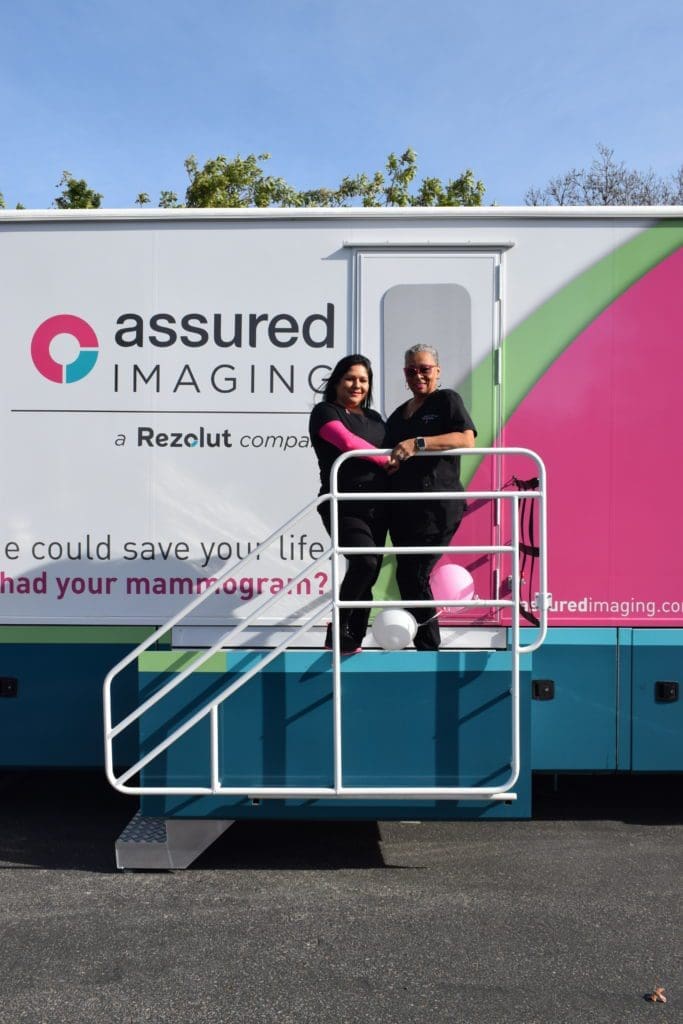 Two people stand smiling on the stairs of a mobile medical imaging unit with Assured Imaging and a Rezolut company written on the side. Pink and white balloons decorate the railing.
