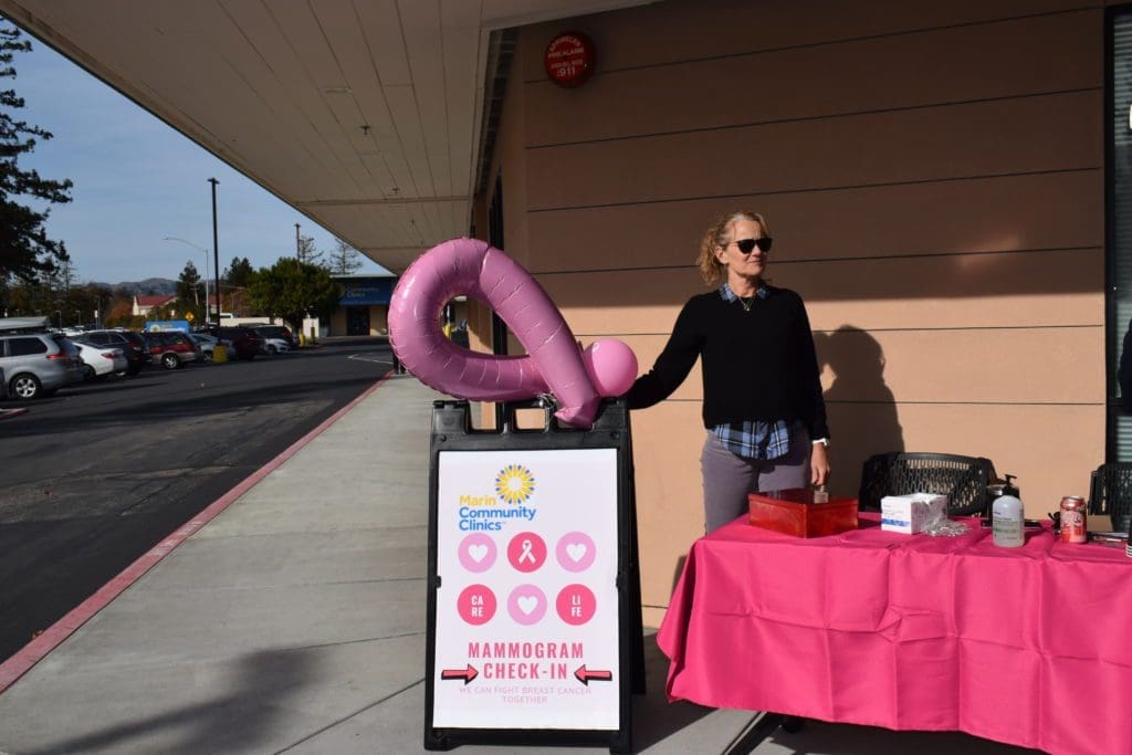 A woman stands next to a pink table and a sign for mammogram check-in, holding a large pink ribbon balloon outside a building on a sunny day.