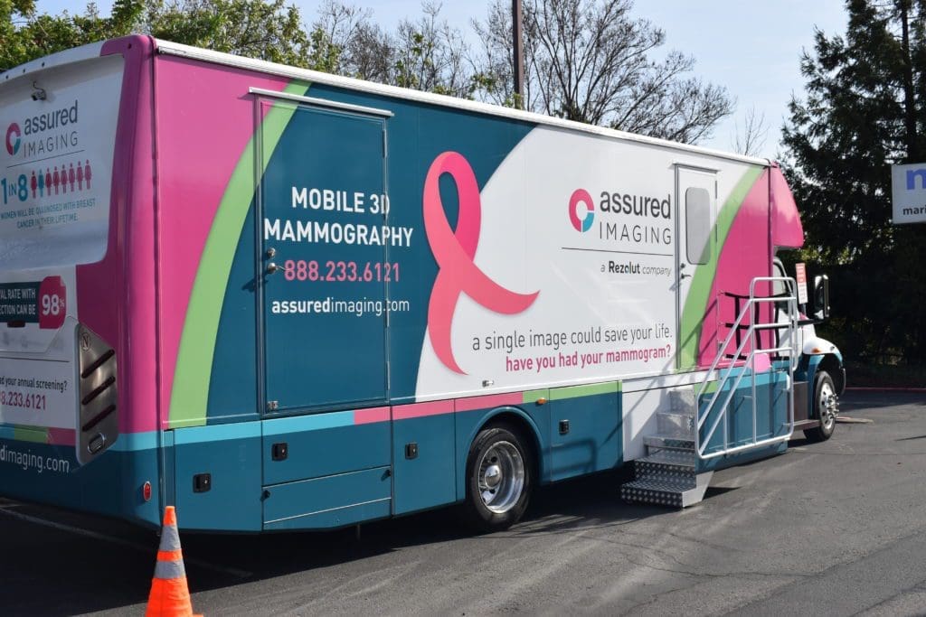 A colorful mobile 3D mammography unit is parked outdoors. The vehicle has a large pink ribbon, contact information, and the slogan “A single image could save your life. Have you had your mammogram?” on its side.