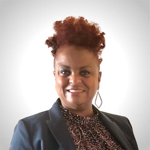 A woman with curly, reddish hair is smiling. She is wearing a dark blazer over a patterned shirt and large, dangling earrings. The background is plain and light-colored.