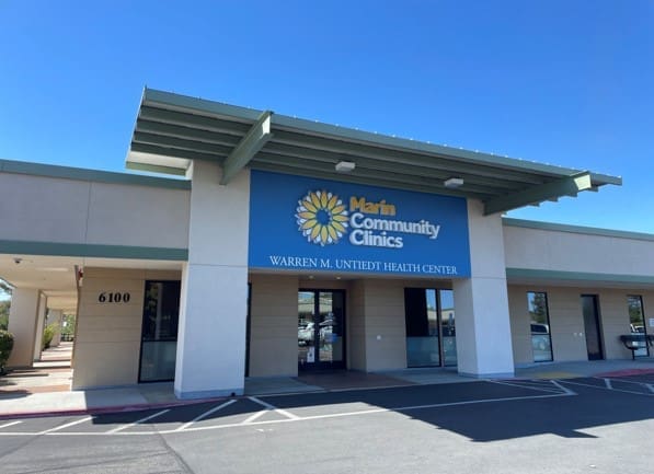 Single-story building with a blue sign reading Marin Community Clinics, Warren M. Unutedt Health Center. The building’s address is 6100. The sky is clear and blue.