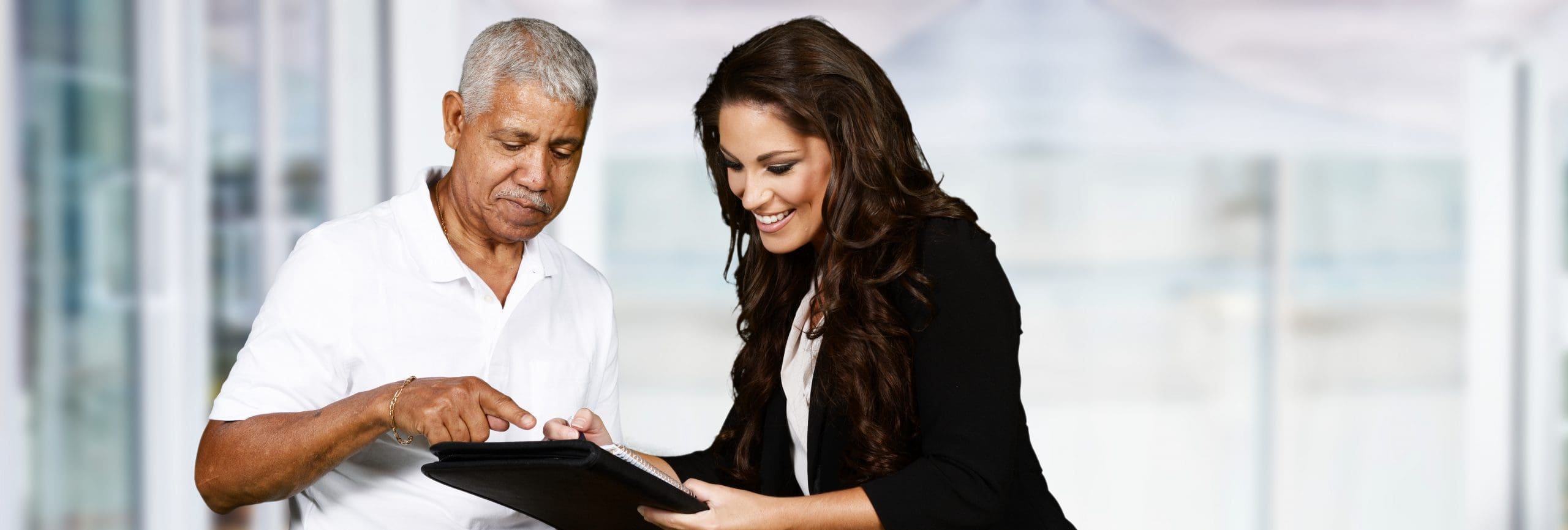 An older man and a younger woman smiling and looking at a tablet together in a bright, modern office setting. The man is pointing at the screen while the woman holds the device and takes notes.