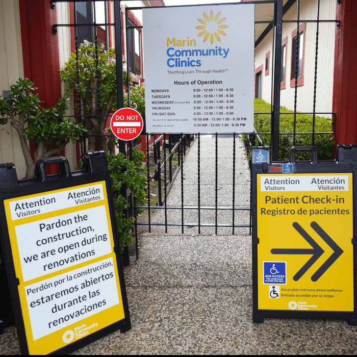 Entrance to Marin Community Clinics with signs about hours, construction notice in English and Spanish, patient check-in instructions, and a “Do Not Enter” sign on the gate. Accessible entrance and masks are required.