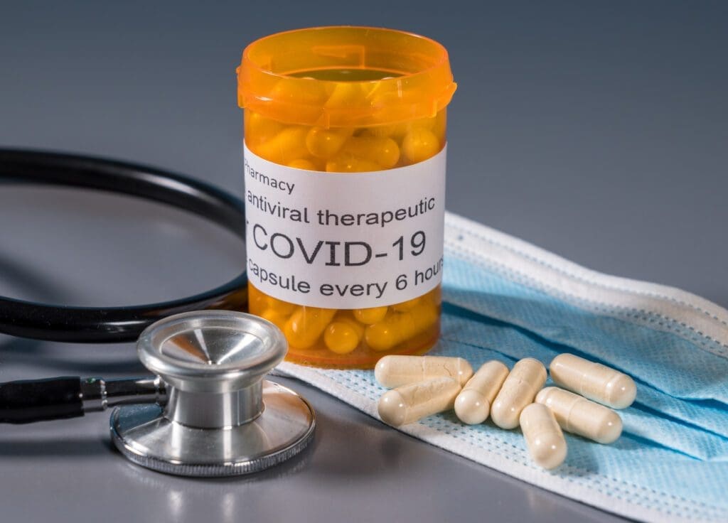 A bottle labeled COVID-19 antiviral therapeutic with yellow capsules, loose capsules, a stethoscope, and a blue surgical mask on a gray surface.