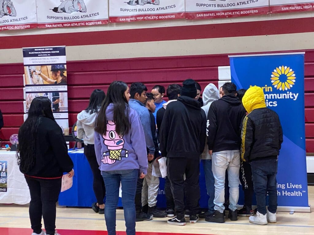 A group of people stand closely together at an indoor event, facing informational booths with banners, including one for Marin Community Clinics. Some people hold papers, and gym bleachers are visible in the background.