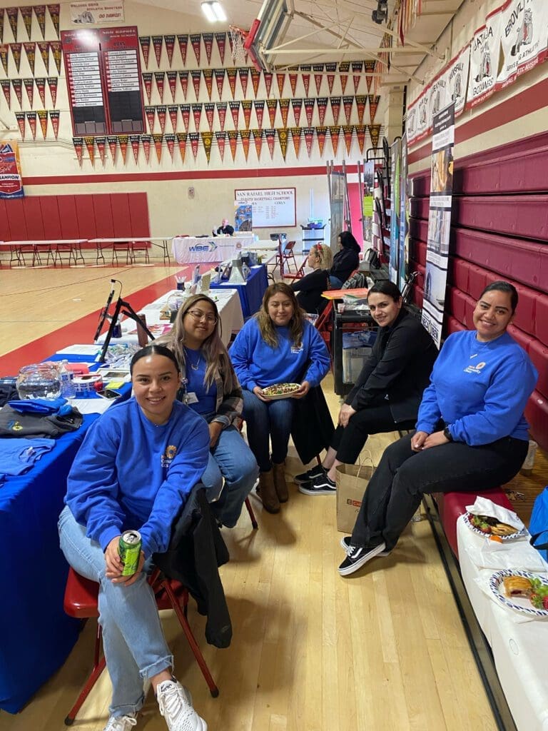 Five women sit and smile at a table in a gymnasium, with informational booths and decorations in the background. Some hold food and drinks, and all wear blue tops. The gym has red walls and banners hanging above.