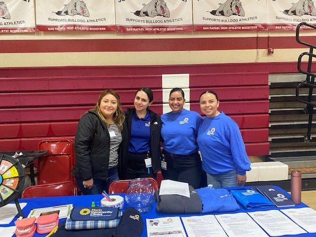 Four women stand smiling behind an information table at a gymnasium, with pamphlets, dental models, and promotional items on the table. Bulldog Athletics banners hang on the wall behind them.