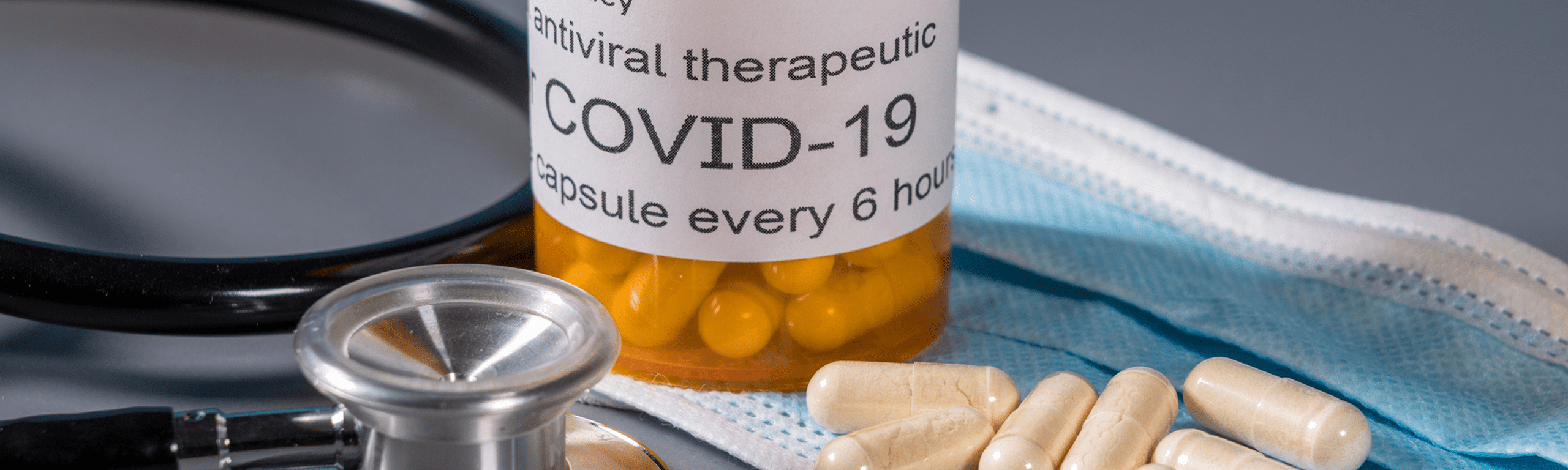 A bottle labeled antiviral therapeutic COVID-19 capsule every 6 hours with yellow capsules, white pills, a stethoscope, and a disposable face mask on a gray surface.