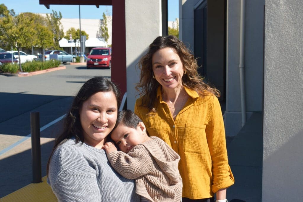 Two women stand outside on a sunny day. One woman, wearing a gray sweater, holds a small child in a brown sweater. The other woman, in a yellow blouse, stands beside them, all smiling at the camera.