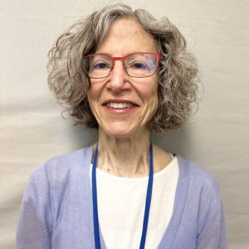 An older woman with curly gray hair, red glasses, and a big smile wearing a white top, light purple cardigan, and a blue lanyard stands in front of a plain, light-colored background.