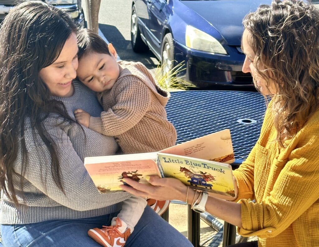 Two women sit at an outdoor table with a young child. One woman holds the child in her arms while the other reads the book Little Blue Truck to them. All are dressed in warm clothes and appear to be enjoying the story.