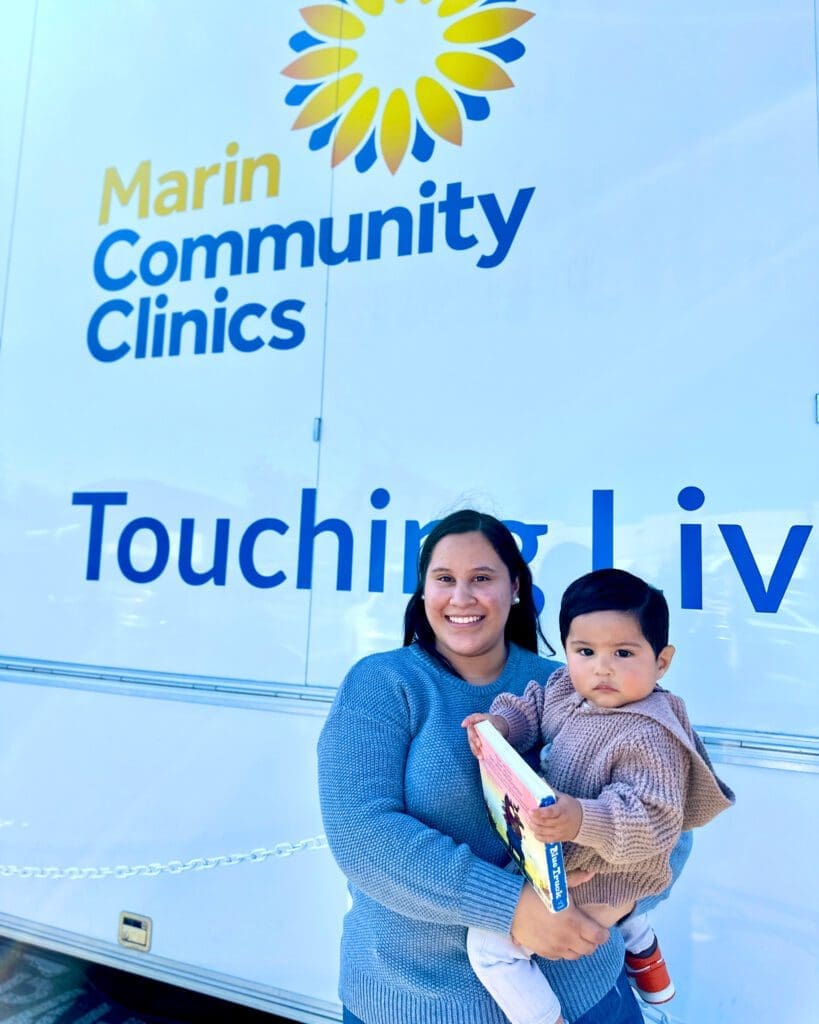 A smiling woman holds a toddler and a book in front of a mobile clinic with the words Marin Community Clinics and Touching Lives visible on the side.