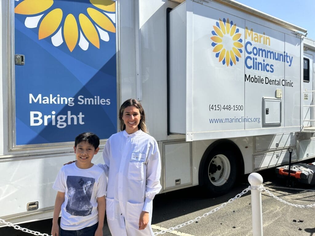 A young boy and a smiling dentist in a white coat stand in front of a Marin Community Clinics Mobile Dental Clinic truck with a bright blue and white sign that reads Making Smiles Bright.