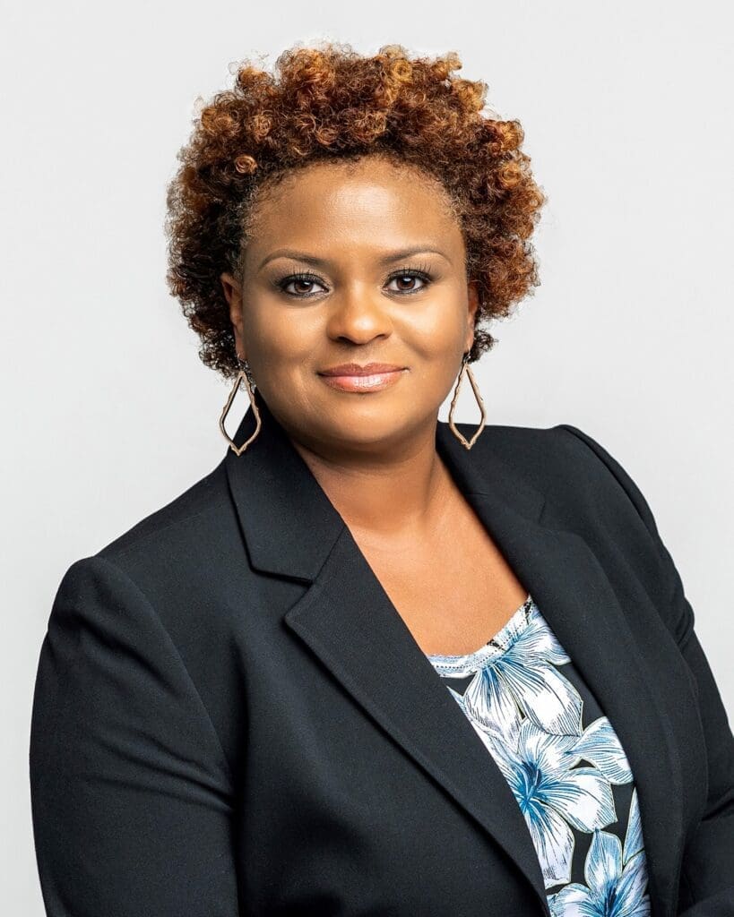 A woman with short curly hair wearing a black blazer, geometric earrings, and a blue-and-white floral top, smiling slightly against a plain light background.