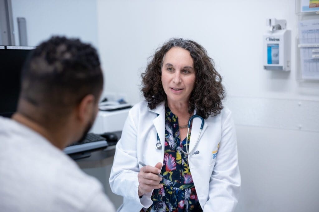 A doctor with curly hair and a stethoscope talks to a patient in a medical office. The patient is seen from behind, and the setting is bright and clinical.