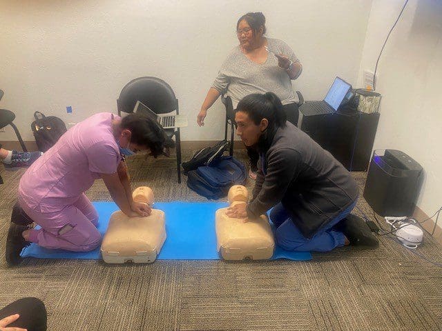 Two people kneel on a blue mat practicing CPR chest compressions on mannequins, while an instructor sits nearby, observing and giving instructions in a classroom setting. Laptops and chairs are visible around them.
