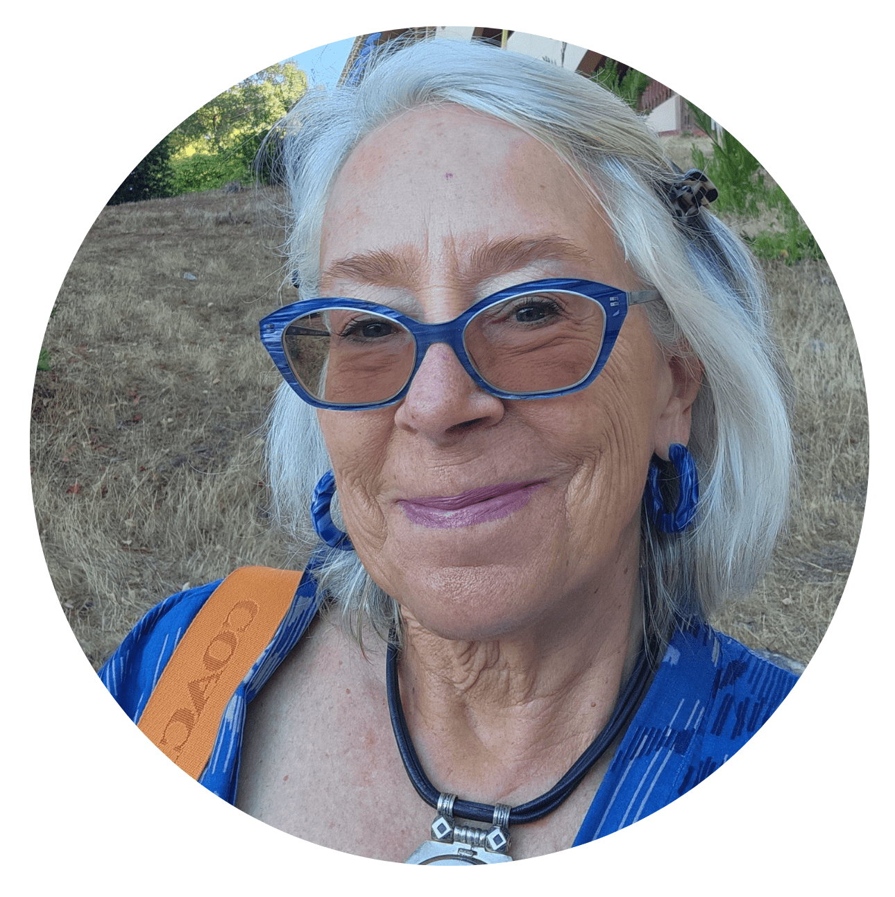 An older woman with white hair, wearing blue glasses and earrings, smiles outdoors. She has a necklace and a Coach bag strap, and is dressed in a blue patterned top with dry grass and greenery in the background.