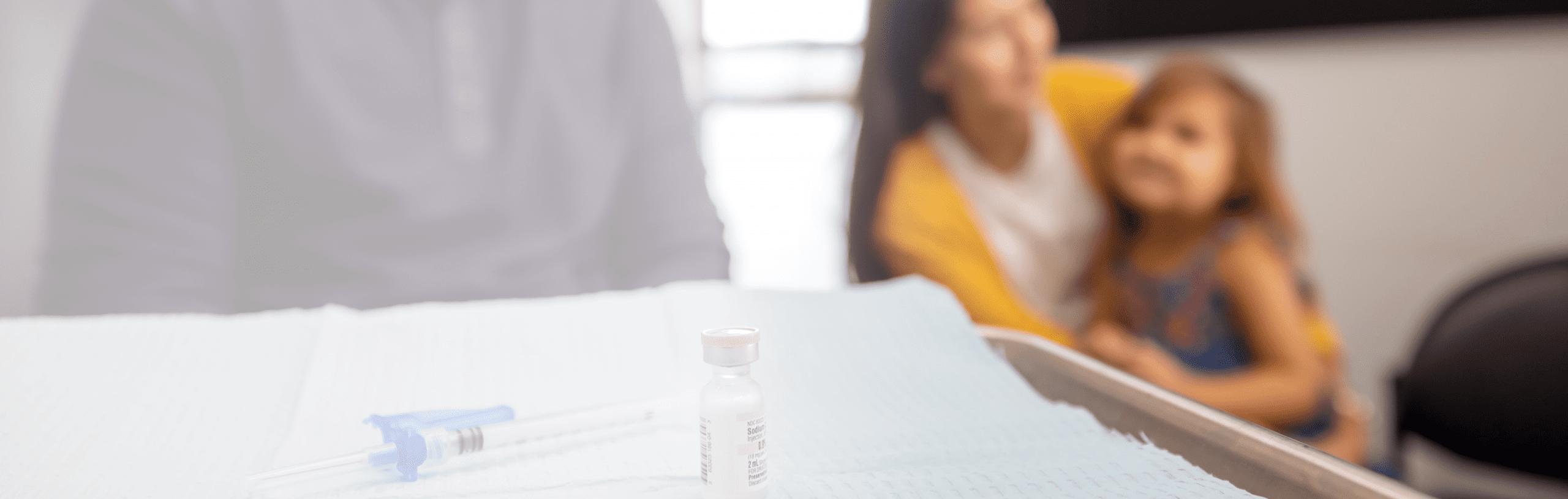 A vial of vaccine and a syringe are on a medical tray in focus, while a woman with a child sitting on her lap are blurred in the background, suggesting a medical or vaccination setting.