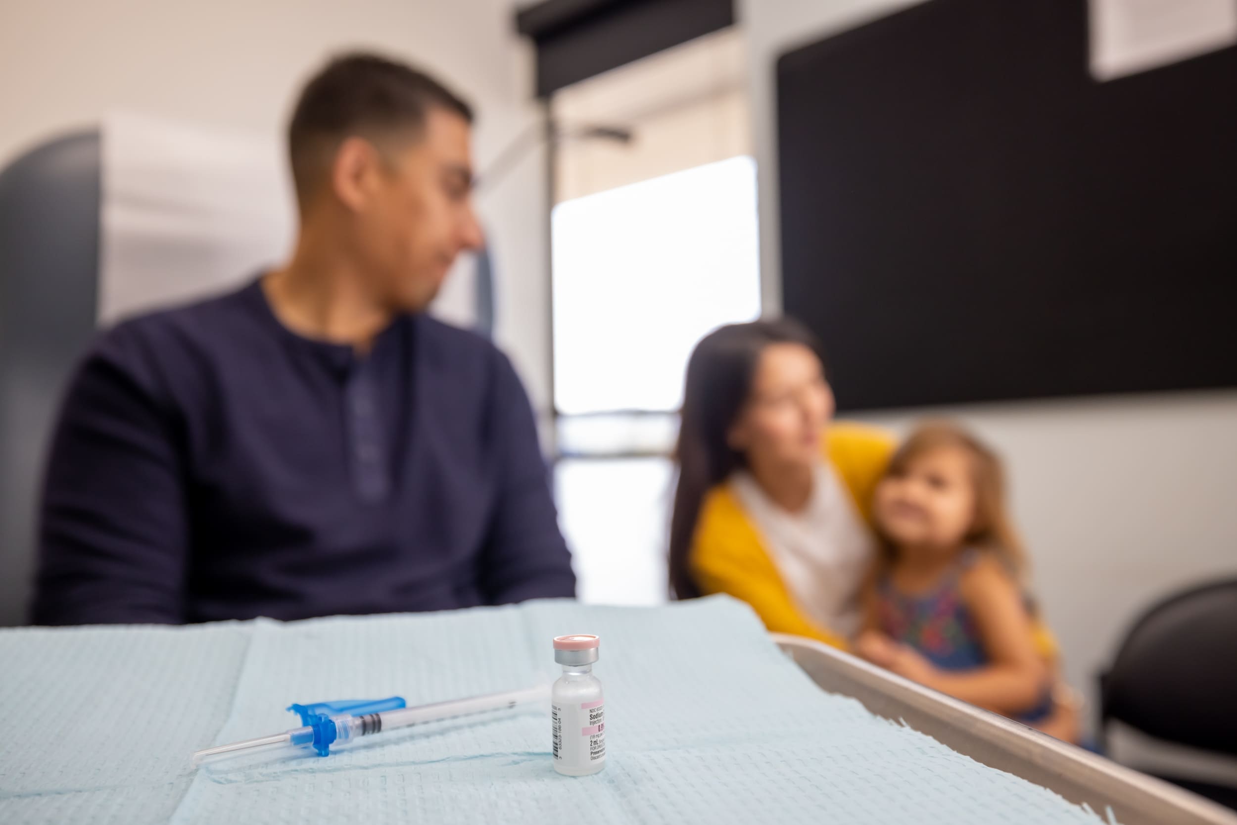 A syringe and a vaccine vial sit on a medical tray in focus, while a blurred adult man, woman, and young girl are visible in the background of a clinic room.