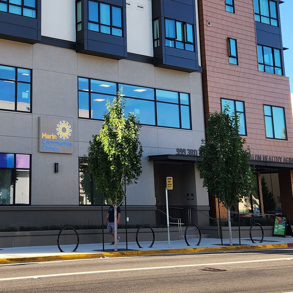 A modern building with “Marin Community Clinics” sign, large windows, young street trees, a sidewalk, a bike rack, and someone walking past. There is a car parked on the street in front.