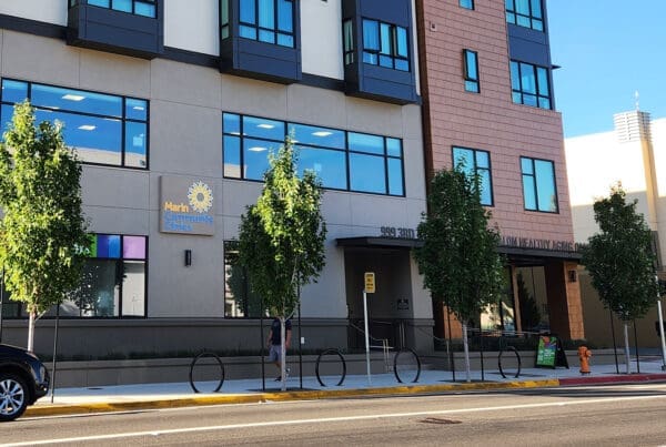 A modern building with “Marin Community Clinics” sign, large windows, young street trees, a sidewalk, a bike rack, and someone walking past. There is a car parked on the street in front.