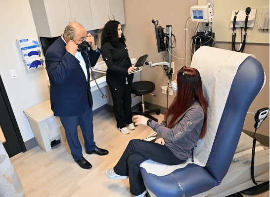A doctor puts on a stethoscope as a nurse stands by a computer, while a patient with long red hair sits on an exam table in a medical exam room.
