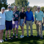 Six adults stand smiling together on a golf course, dressed in casual golf attire. Trees, grass, and golfers are visible in the background on a sunny day.
