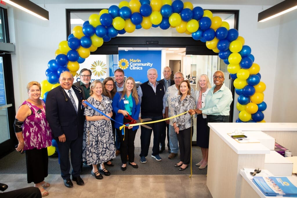 A group of people smile and pose as they cut a yellow ribbon under a blue and yellow balloon arch at the entrance of Marin Community Clinics. A Marin Community Clinics sign is displayed in the background.