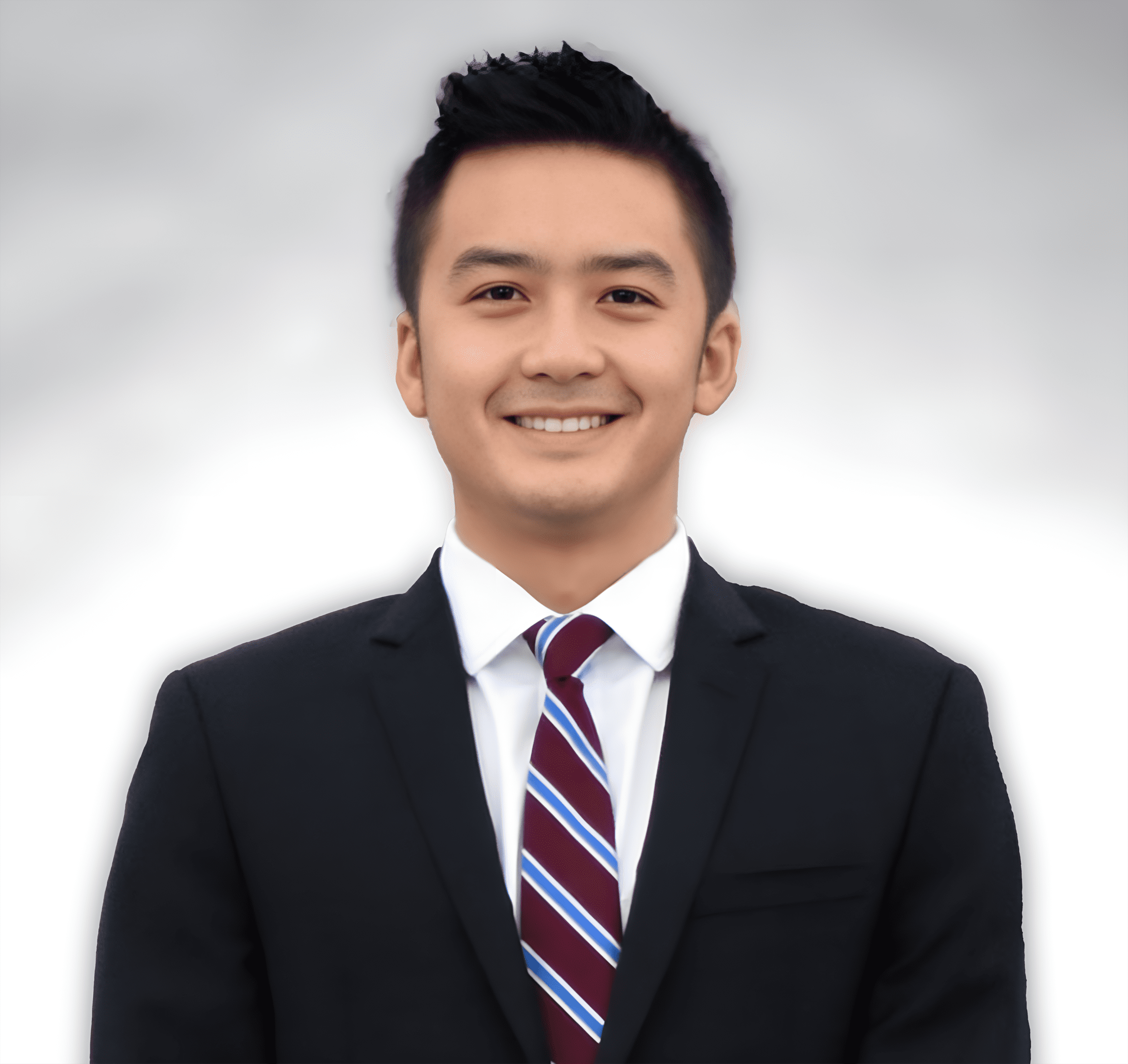 A young man with short black hair smiles while wearing a black suit, white shirt, and striped tie, standing against a blurred gray and white background.
