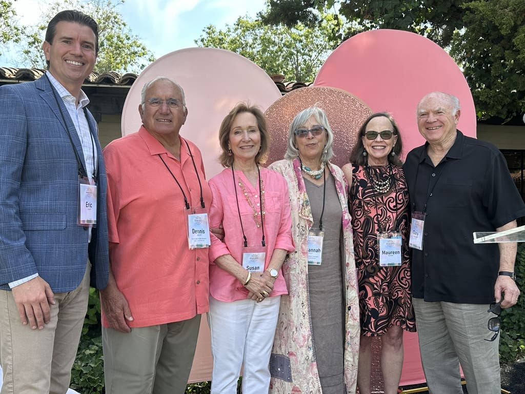 Six smiling adults pose outdoors in front of large pink circular decorations. They are dressed in colorful, semi-formal attire and all wear name tags. Trees and a building are visible in the background.