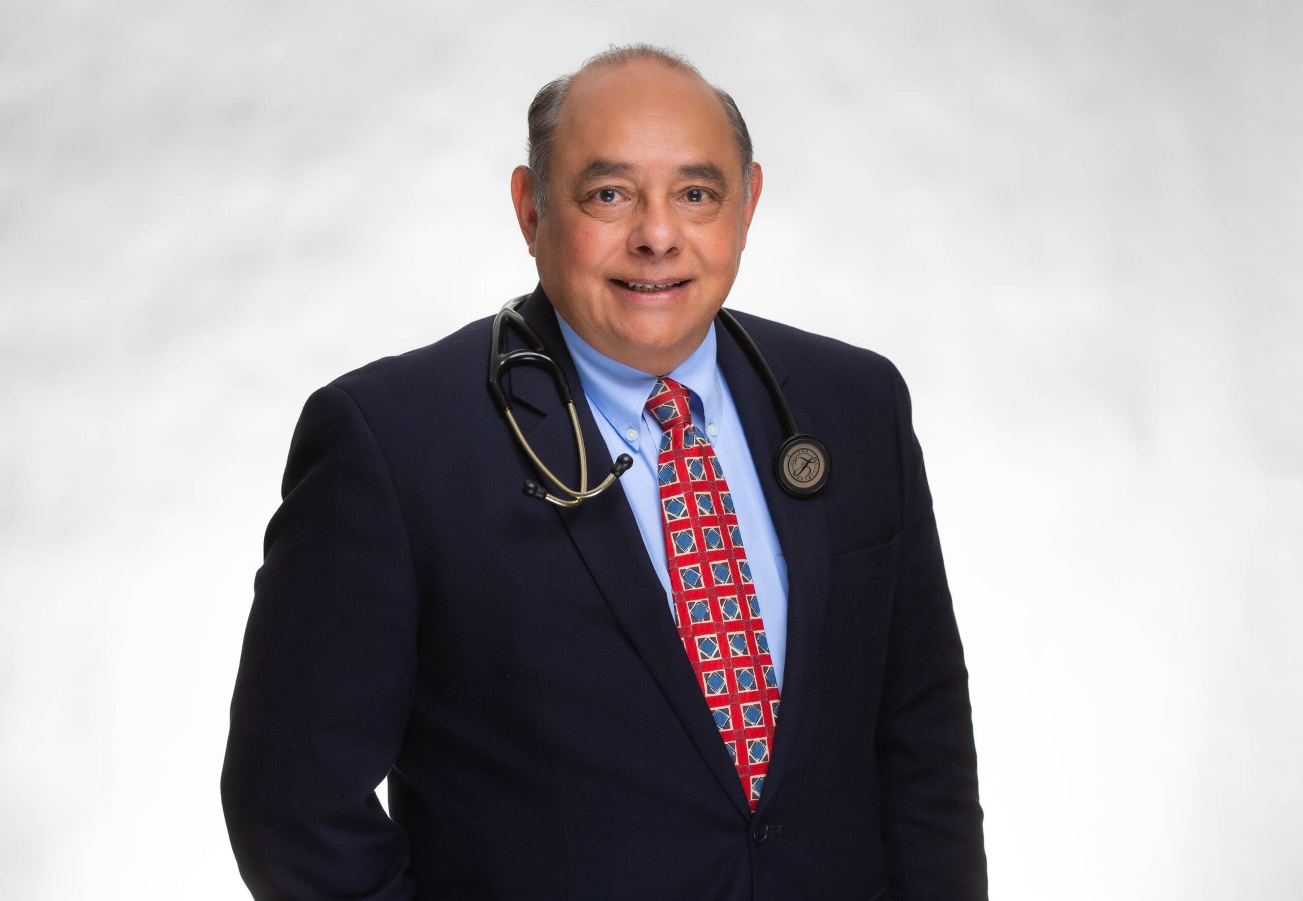 A smiling man in a suit, blue shirt, and colorful patterned tie stands against a white background with a stethoscope draped around his neck.