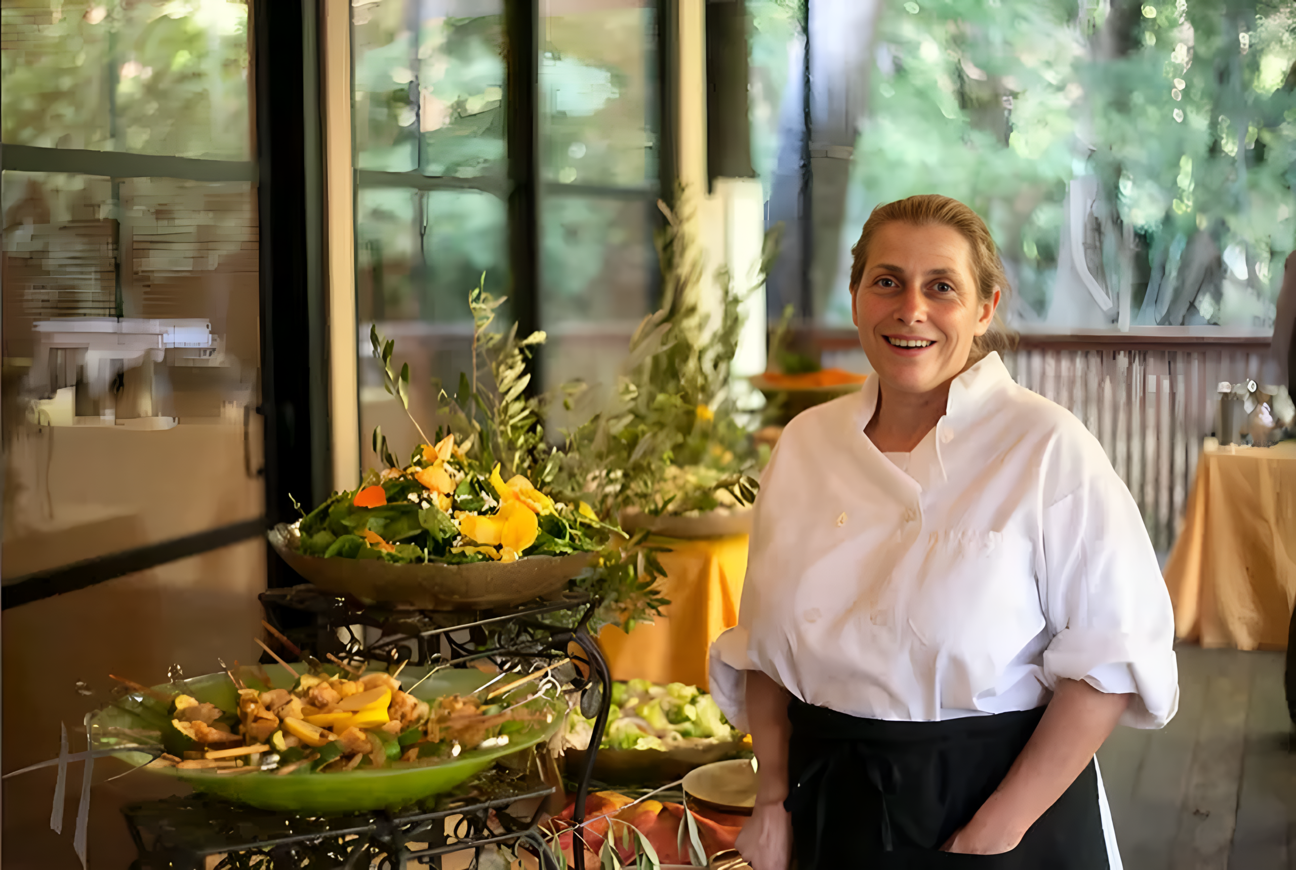 A chef in a white coat stands smiling beside a table of colorful salads and fresh greens, with large windows and trees visible in the background.