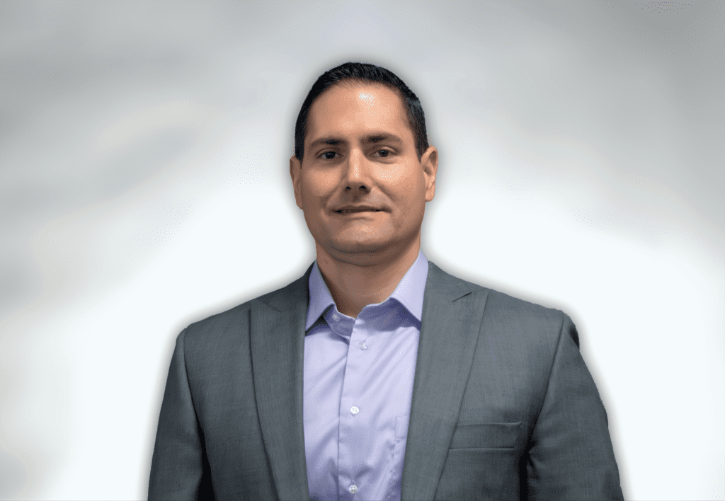 A man with short dark hair wearing a gray suit jacket over a light purple dress shirt stands against a plain, light gray background, looking at the camera with a neutral expression.