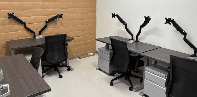 A small office with four desks, each equipped with a black mesh chair, gray drawers, and dual monitor arms. The room has beige and white walls and a clean, minimalist appearance.