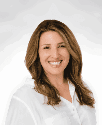 A woman with long, light brown hair and a bright smile wears a white shirt and is standing against a plain, light background.